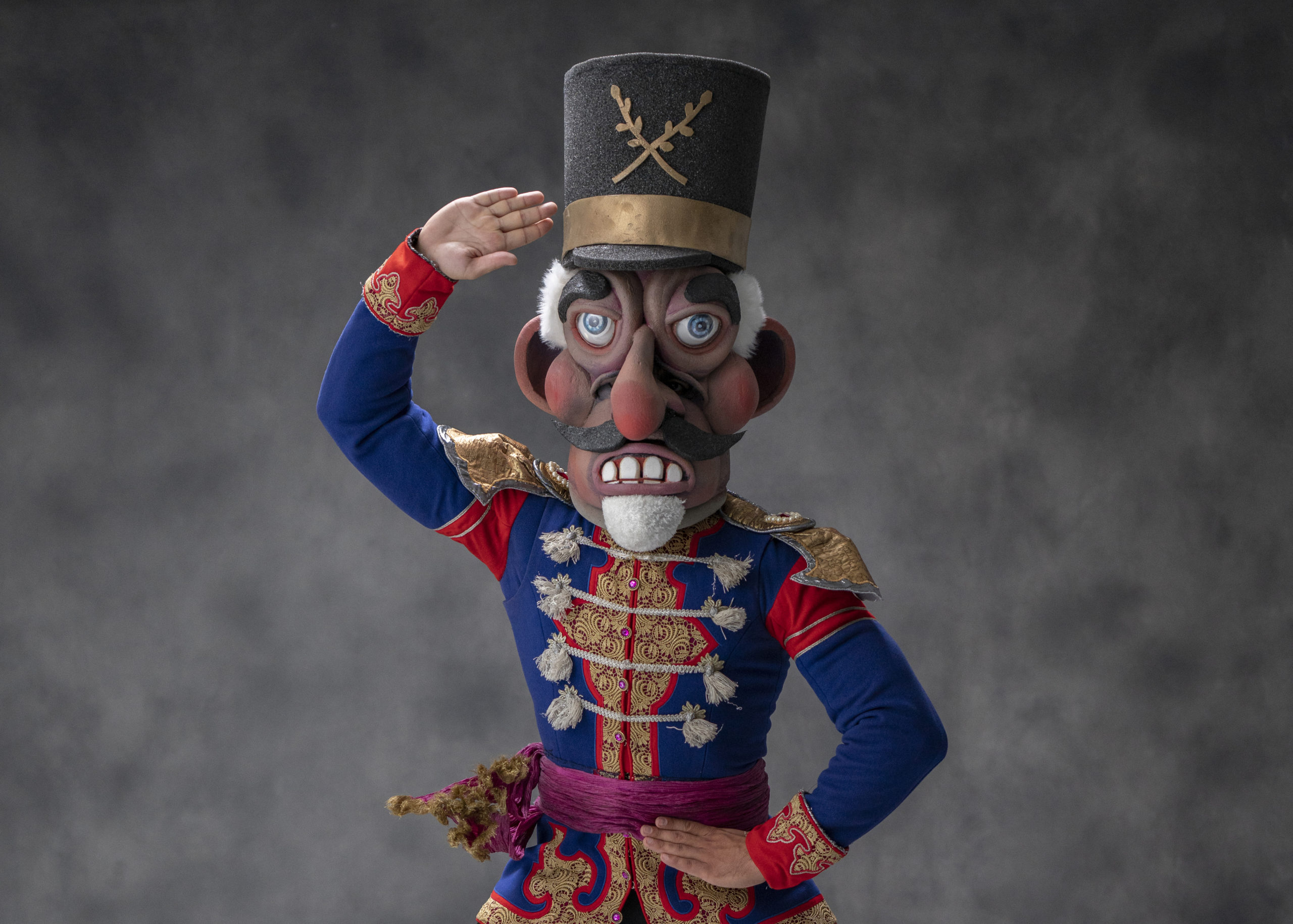 A male dancer is shown from the waist up wearing a Nutcracker costume and large Nutcracker head. He holds his left hand at his waist and salutes with his right hand.