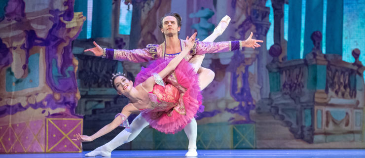 Dancer Andrew Taft, in a purple jacket and white tights, lunges deeply on his left leg and holds his arms out wide while her partner, ballerina Elizabeth Barreto, has her right leg wrapped around his waist while she holds her upper body up in a "fish" lift. She wears a bright pink tutu and tiara, and they dance onstage during a performance of the Nutcracker ballet with backdrops depicting the Land of Sweets.