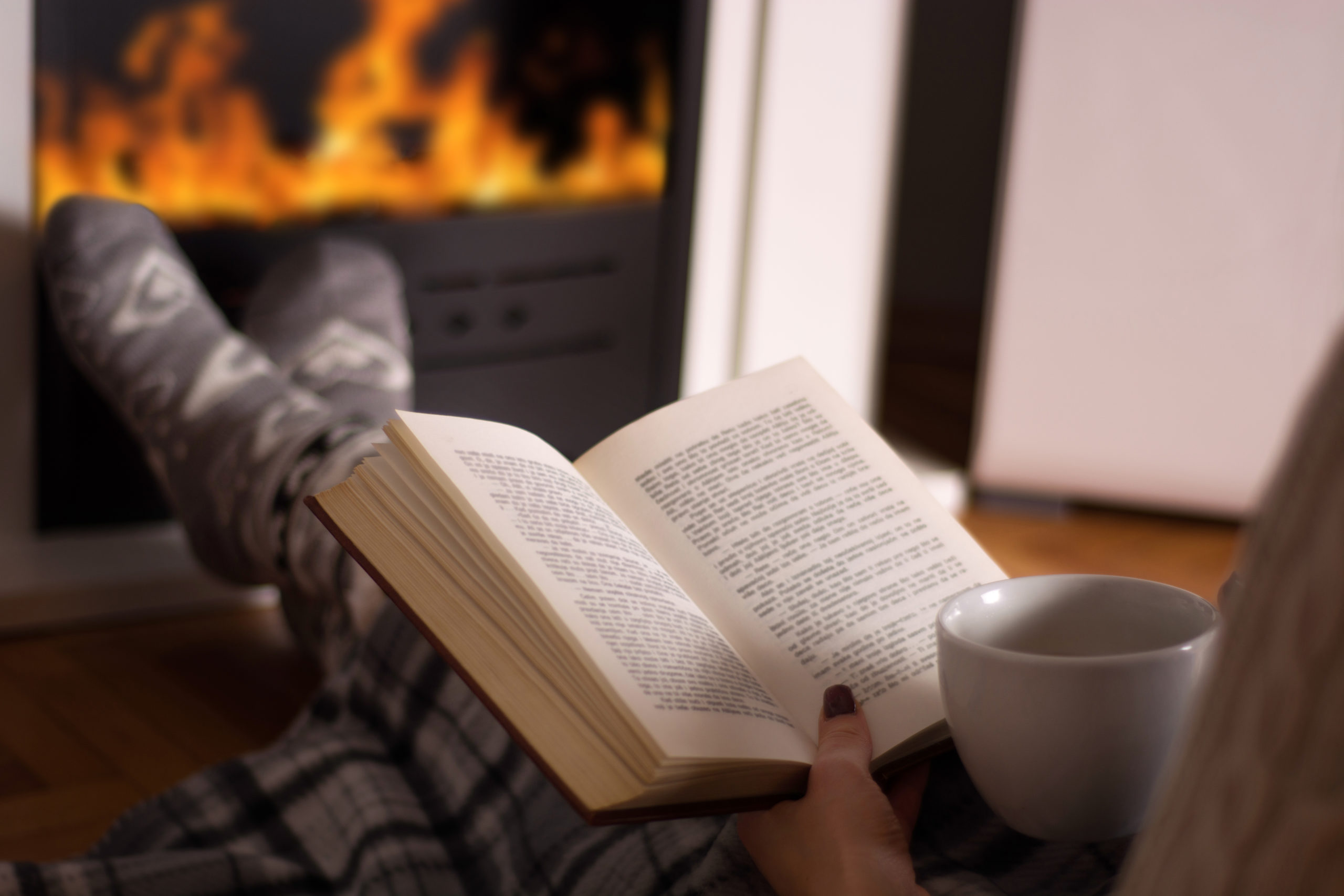 A woman is shown fro the waist down under a plaid blanket, propping her feet on an ottoman and wearing thick wooly socks. She is reading a book and holding a cup of tea while warming her feet in front of the fireplace at home.