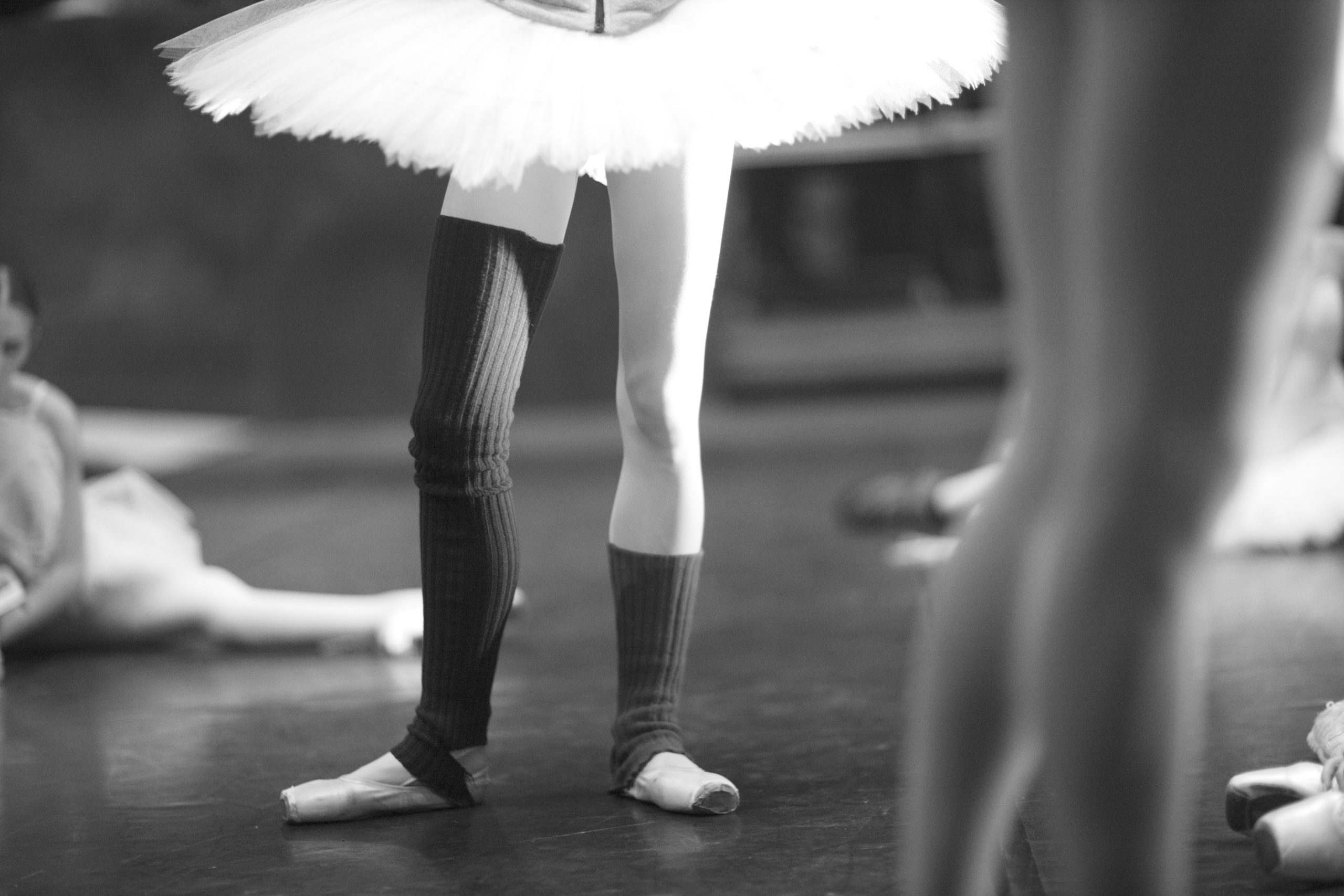 A black-and-white image of a dancer in a tutu, pointe shoes and leg warmers onstage. Blurred dancers warm up around her.