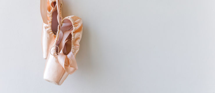 A pair of satin pink pointe shoes hang against a blank wall
