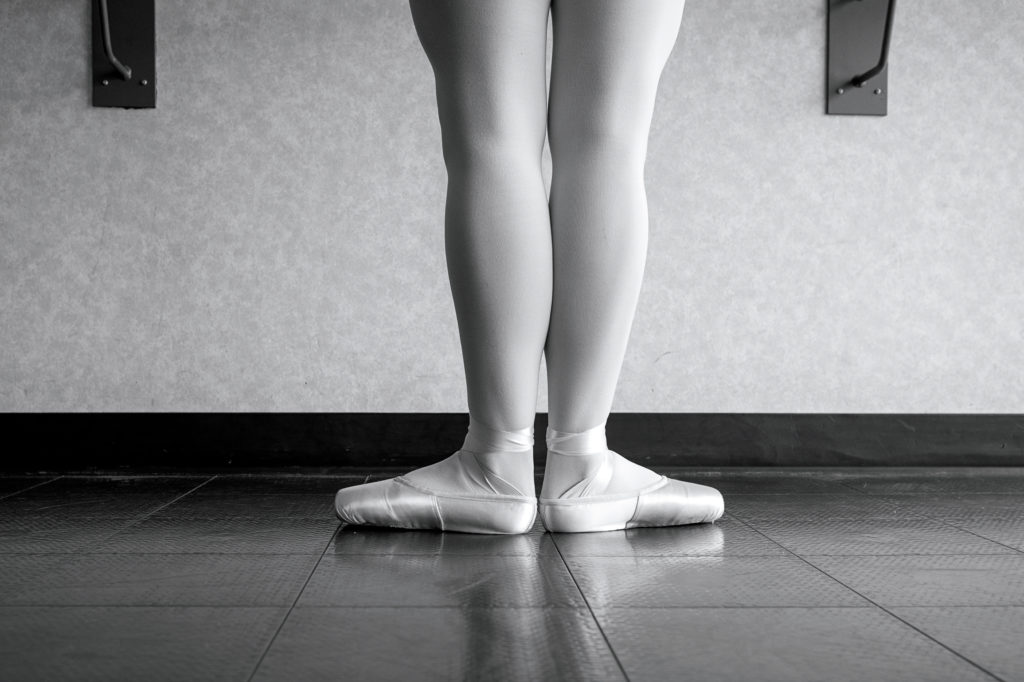 In this black and white image, a female dancer is shown from the thighs down standing in first position. She wears tights and pointe shoes.
