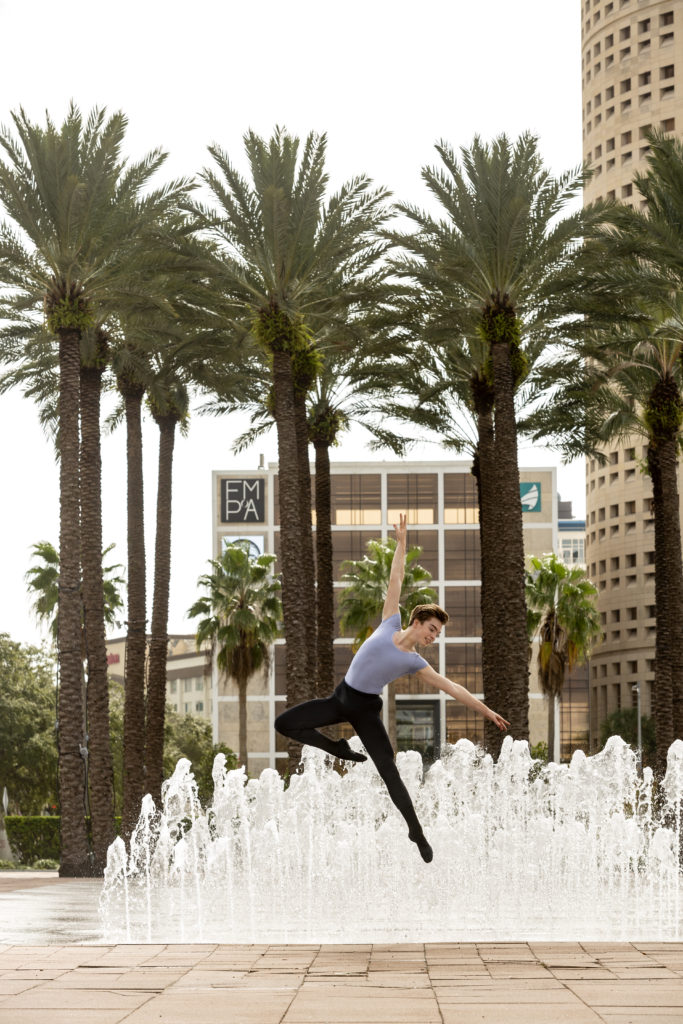 A teen male ballet dancer photographed midair, with his left leg extended and right leg in a high passé. He is dancing in a plaza with fountains and palm trees behind him.