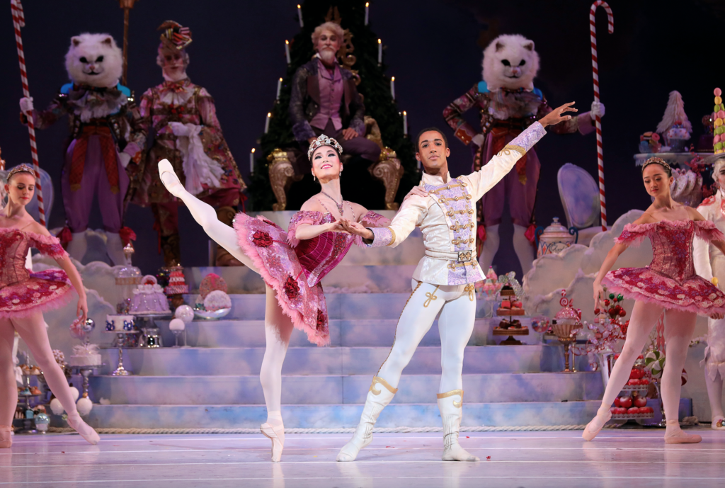 Soo Youn Cho, in a dark pink tutu with floral trim and a large crown, poses in attitude on pointe and holds Harper Watters' right hand and shoulder for balance. Watters wears a white jacket, tights and dance boots with lavender and gold trim and stands with his wieght on his left foot and his left arm out the side. Behind them onstage, a cast of characters from the Nutcracker pose and watch.