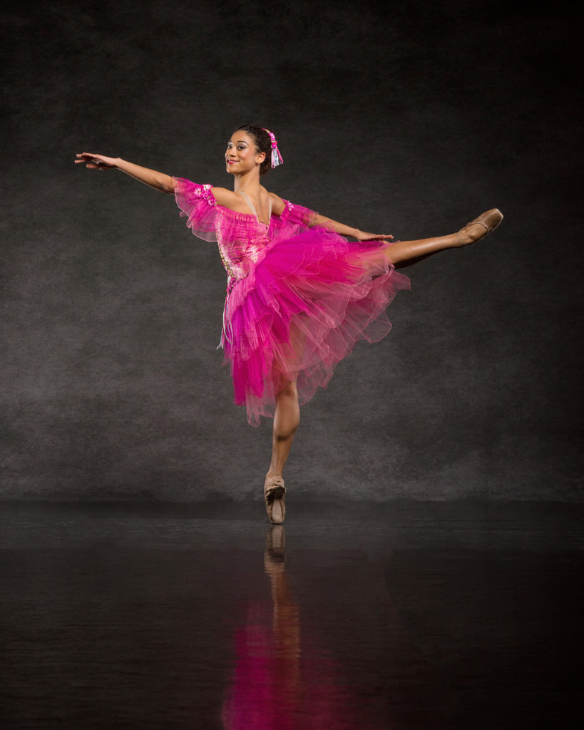 Princess Reid, wearing a bright pink Romantic tutu, poses on pointe in a second arabesque with her left leg raised. She looks out towards the camera with a small smile.