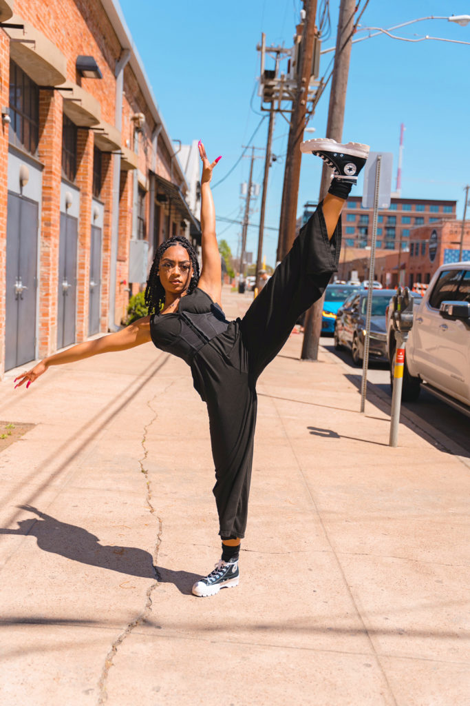 Caeli Blake, in a black jumpsuit, stands on a city sidewalk and kicks her left leg up, foot flexed, while leaning slightly back with her upper body. She wears black Converse tennis shoes and poses her left arm up high and her right arm out t the side.