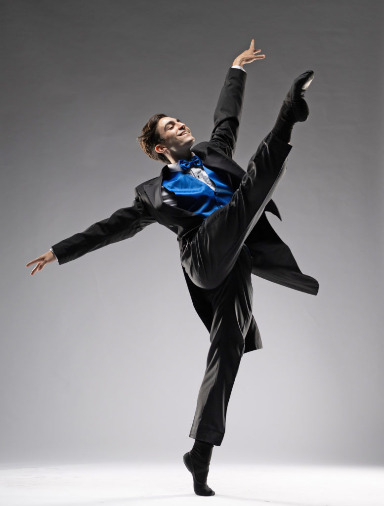 Ballet dancer Jaysan Stinnett performs a développé croisé devant with her right leg, his left foot on relevé. Wearing a black tuxedo with bright blue vest and bow tie, black socks and black ballet slippers, he lifts his left arm high and opens his right arm to the side.