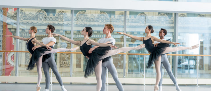 Three women balance in arabesque on pointe while their male partners support them.