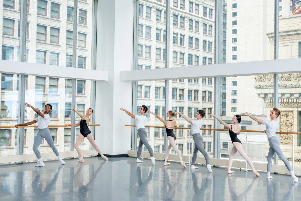 Young adult dancers at the barre in tendu derrière in a studio with floor-to-ceiling glass walls.