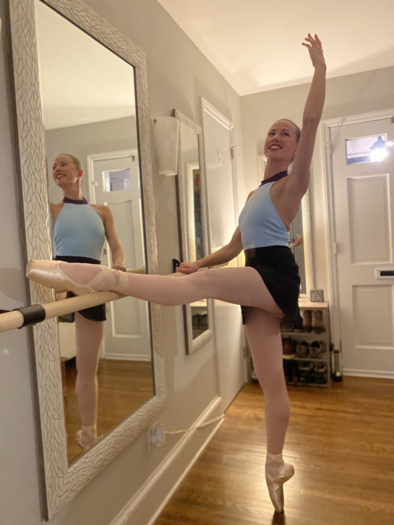 Katherine Nobles stands at a ballet barre inside her home and rests her left leg on the barre in croise devant while balancing on pointe on her right foot. She holds the barre with her right hand and wears a blue leotard, black skirt, and pink tights and pointe shoes.