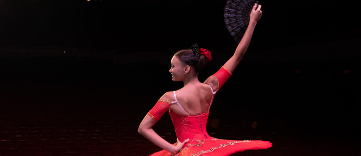 A teen female ballet dancer kneels onstage, while wearing pointe shoes and a red tutu and holding a fan. The bright theater lights illuminate her and the stage.