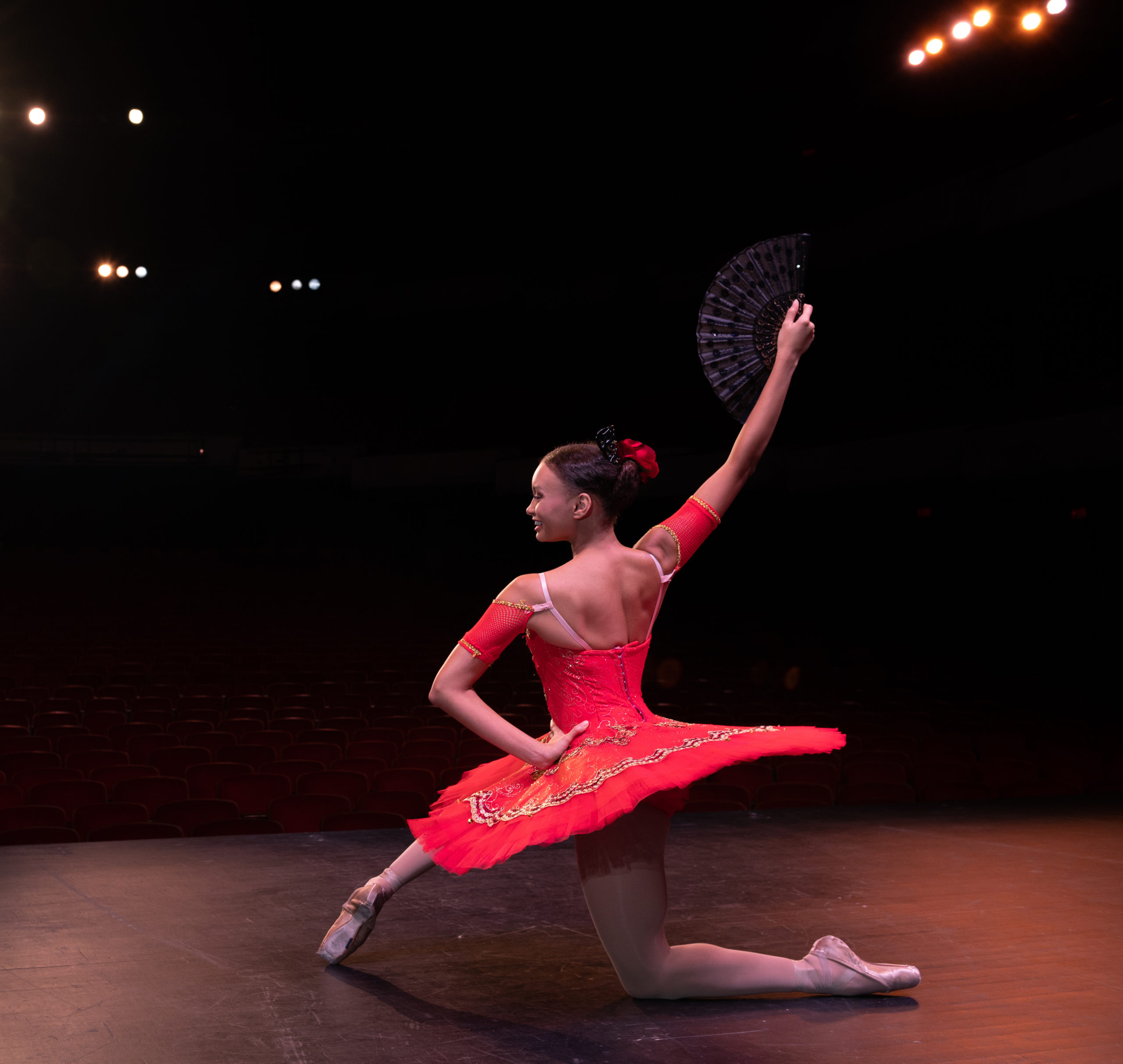 A teen female ballet dancer kneels onstage, while wearing pointe shoes and a red tutu and holding a fan. The bright theater lights illuminate her and the stage.