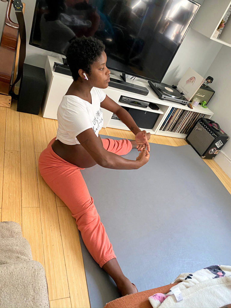 Ingrid Silva, who is pregnant, sits on the floor of her living room in a straddle stretch holding her arms in first position. She wears a white crop top, exposing her belly, and pink sweat pants.