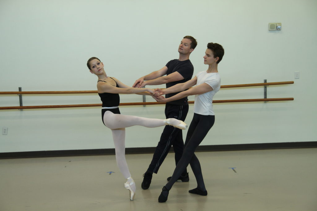 Peter Start stands between a male and female student as they practice a pose together. They are in front of the barre in a dance studio.
