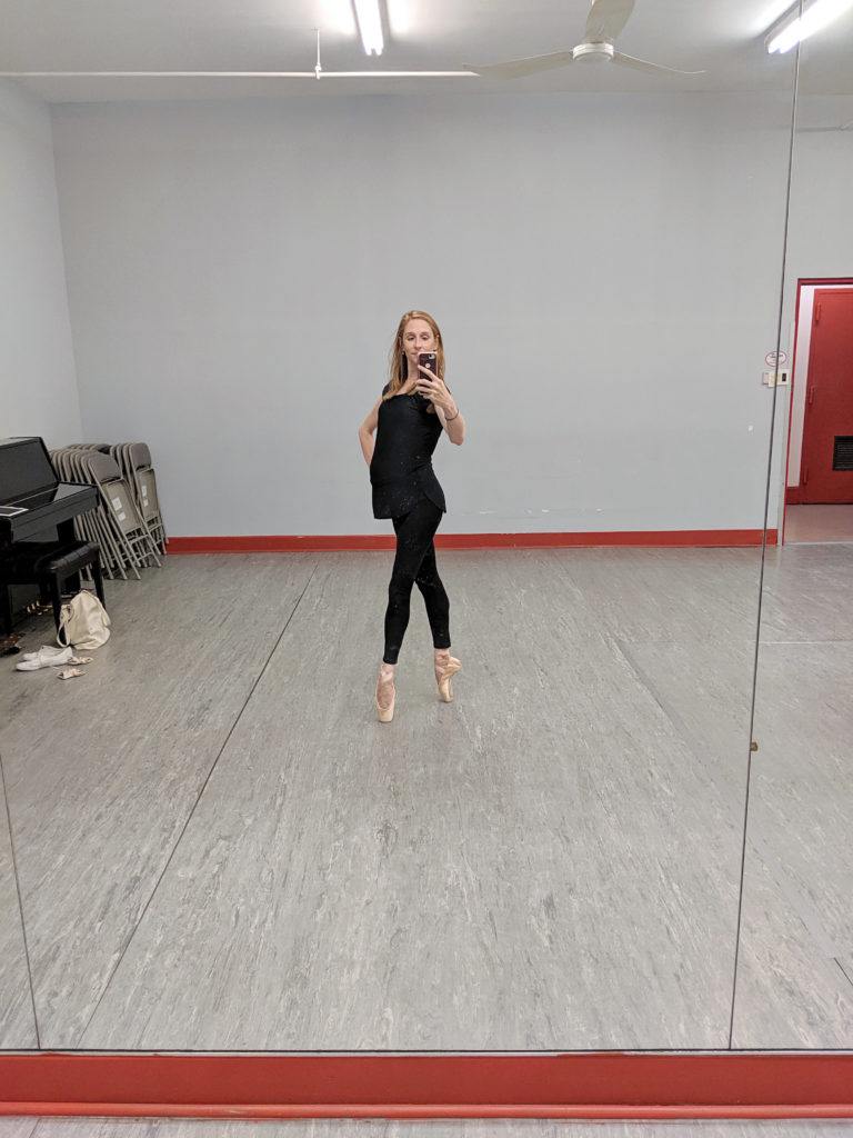 Ashley Laracey poses in fourth position on pointe with her right hand on her hip and takes a selfie in the mirror. She is pregnant and wears a short black jumper dress and black tights..