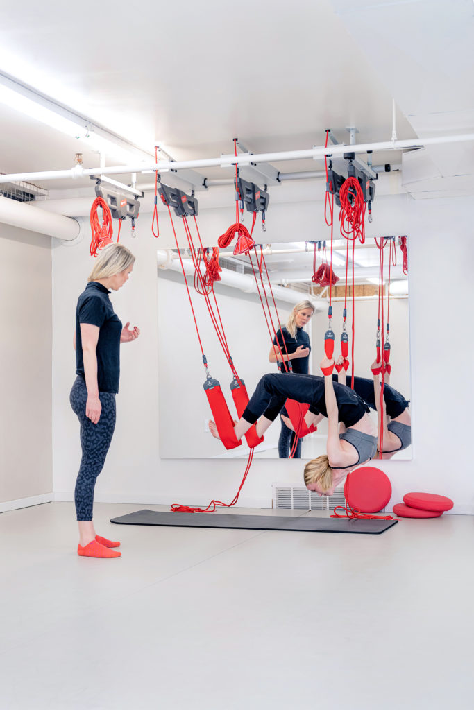 Nikki Bybee, wearing dark athleisure clothing, stands to the left and watches a young female dancer move on a Redcord trainer. The dancer, in a sports bra and leggings, is suspended in the air by a series of red straps and ropes connected to the ceiling, and leans her upper body backwards so that her face faces the floor.