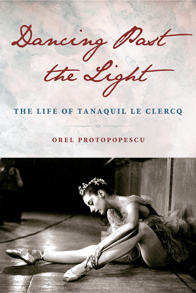 The photo shows the book cover of "Dancing Past the Light: The Life of Tanaquil Leclerq" by Orel Protopopescu. The book title is shown in the top half of the cover in cursive lettering. A black and white photo of Tanaquil LeLerq in costume, sitting on the floor backstage and adjusting her pointe shoes, is on the bottom half.
