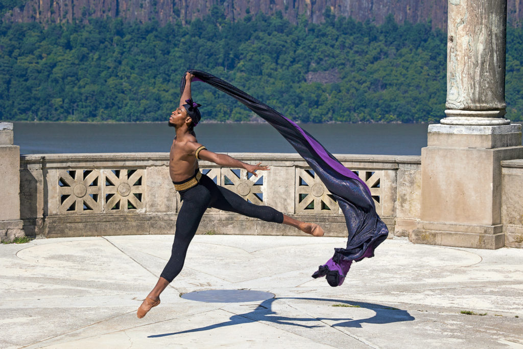 Khayr Muhammad, wearing dark tights with a natrual fiber belt and dark durag headpiece performs a temps levé and holds a long purple cape up with his right hand. He dances on a grand marble terrace with large pillars, overlooking a large cliff with river below.