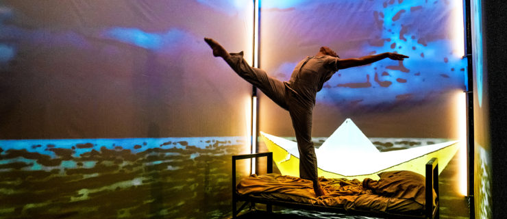 Elijah Lancaster, wearing a prison jumpsuit, performs a dramatic layout on relevé on the mattress of a small twin bed. Projections of the sea and sky surrounding him on the walls of the narrow detention cell set.