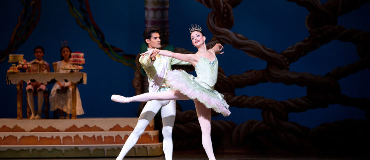 Jennifer Lauren, in a mint green and pink tutu, does a relevé arabesque on pointe with her arms in second. Her partner, Renan Cerdeiro, wears a mint green tunic and white tights, stands behind her and holds both of her hands They dance onstage during a performance of the Nutcracker, and two small children sit behind them in costume in front of a table of sweets.