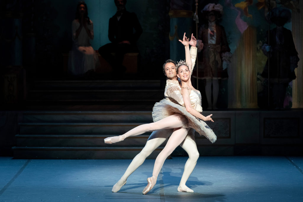 During a performance of The Nutcracker, Paolo Arrais lunges in fourth behind ballerina Viktorina Kapitonova and wraps his right arm around her waist as she performs a low arabesque. She wears a light-colored tutu and tiara, pink tights and pointe shoes and he wears a matching colored jacket, tights and slippers.
