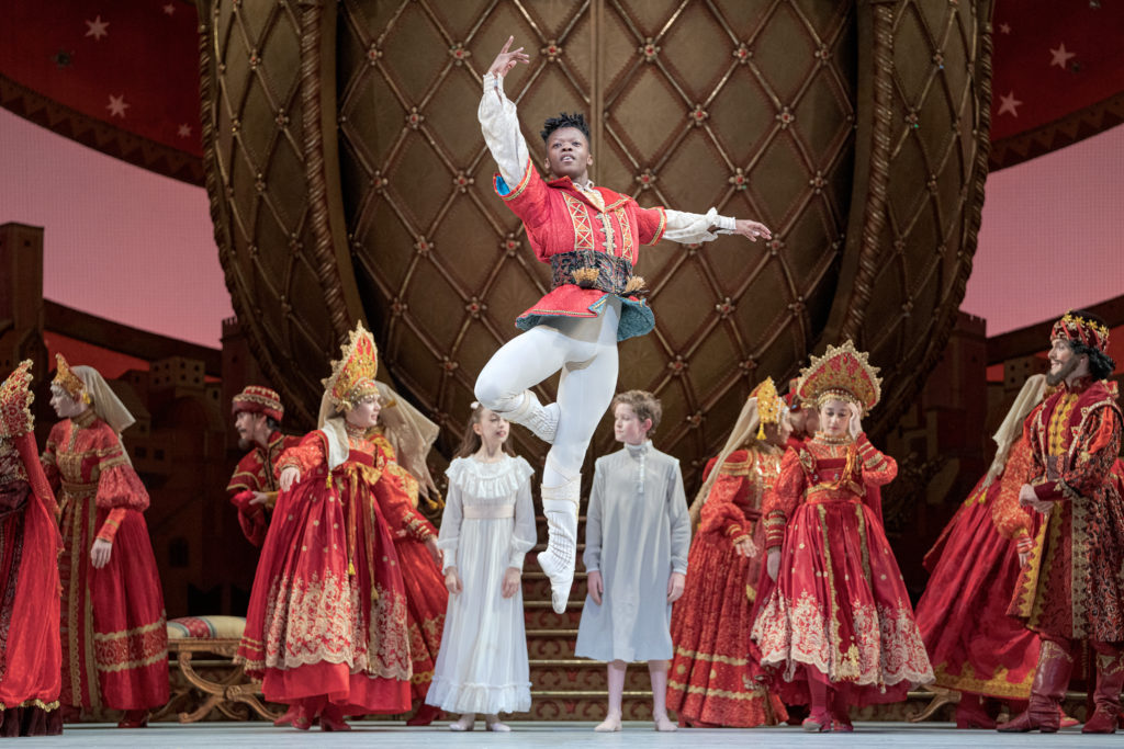 Siphesihle November, costumed as a prince in a red and gold jacket, white tights and white boots, jumps straight up with his right leg in retiré and his arms in third position. Behind him onstage is a boy and girl costumed in nightgowns and a group of dancers in Russian-style costumes.