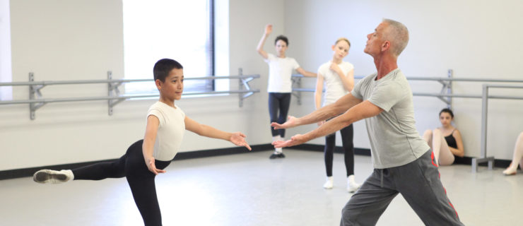 Bill Piner, wearing a light gray T-shirt and dark gray sweatpants coaches a young boy during a ballet class. He stands in front of the boy and stretches both arms out. The dancer, wearing a white t-shirt, black tights and white socks and ballet slippers, makes a similar position with his arms and balances in an attitude with his left leg back.