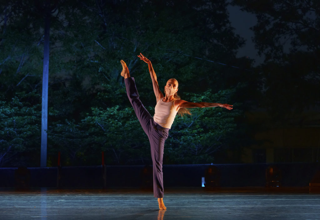 Brooke Gilliam, in purple pants and a tank top, balances on relevé with her right leg extended à la seconde. She is dancing on an outdoor stage at night with trees in the background.