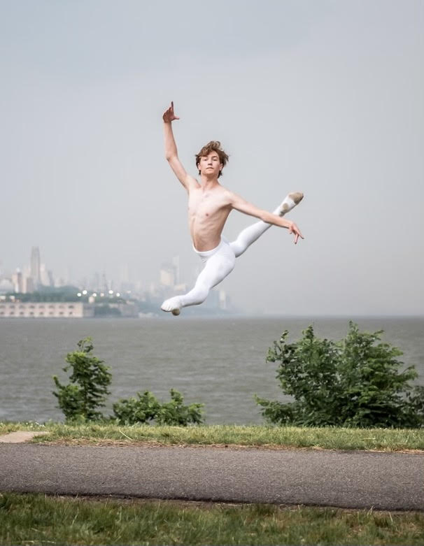 Chase Hanson is jumps in a sissone en avant with his arms in fourth position. He wears white tights and no shirt and behind him is a large, foggy body of water