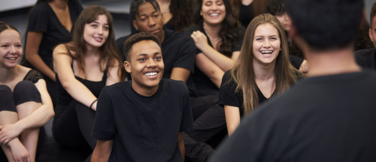 Teacher At Performing Arts School Talking To Students Sitting On Floor In Rehearsal Studio