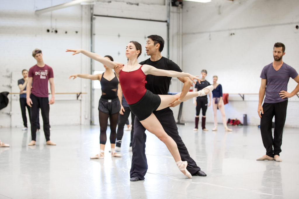 Edwaard Liang holds dancer Grace-Anne Powers by the left thigh and right armpit as she leans forward in arabesque and talking to and demonstrating for the dancers surrounding him. powers wears a red leotard, black booty shorts and pointe shoes, while Liang is dressed in black warm-up pants and a T-shirt. The other dancers, in dancewear, look on in the large studio.
