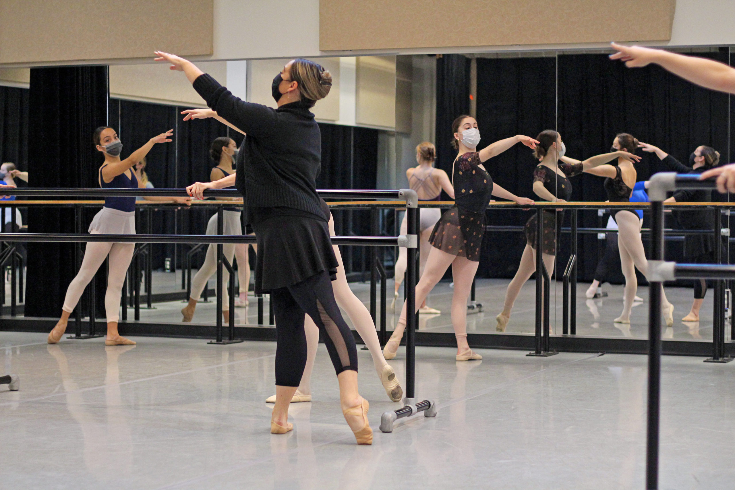 Jennifer Guy Metcalf demonstrates a tendu derriere with her left leg at the barre while her class of dance students follow suit. She wears a face mask, black sweater, black ballet skirt and leggings, and tan ballet slippers. Her students wear also wear face masks, leotards, tights and skirts and ballet slippers in various colors.