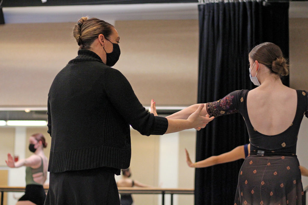 Jennifer Guy Melcalf adjusts the elbow of a college dance student from behind. She wears a black sweater and face mask, while teh dancer wears a long-sleeved leotard with a diamond and floral pattern and matching skirt.