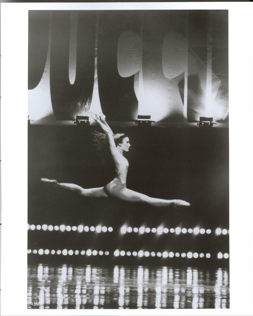 In this black and white photo, Jodie Gates leaps through the air onstage with her arms thrown up above her head. She wears a unitard, her long hair flying free. Behind her, two rows of stage lights glow at the stage's edge, wit a large billboard reading "Pucci" above her.