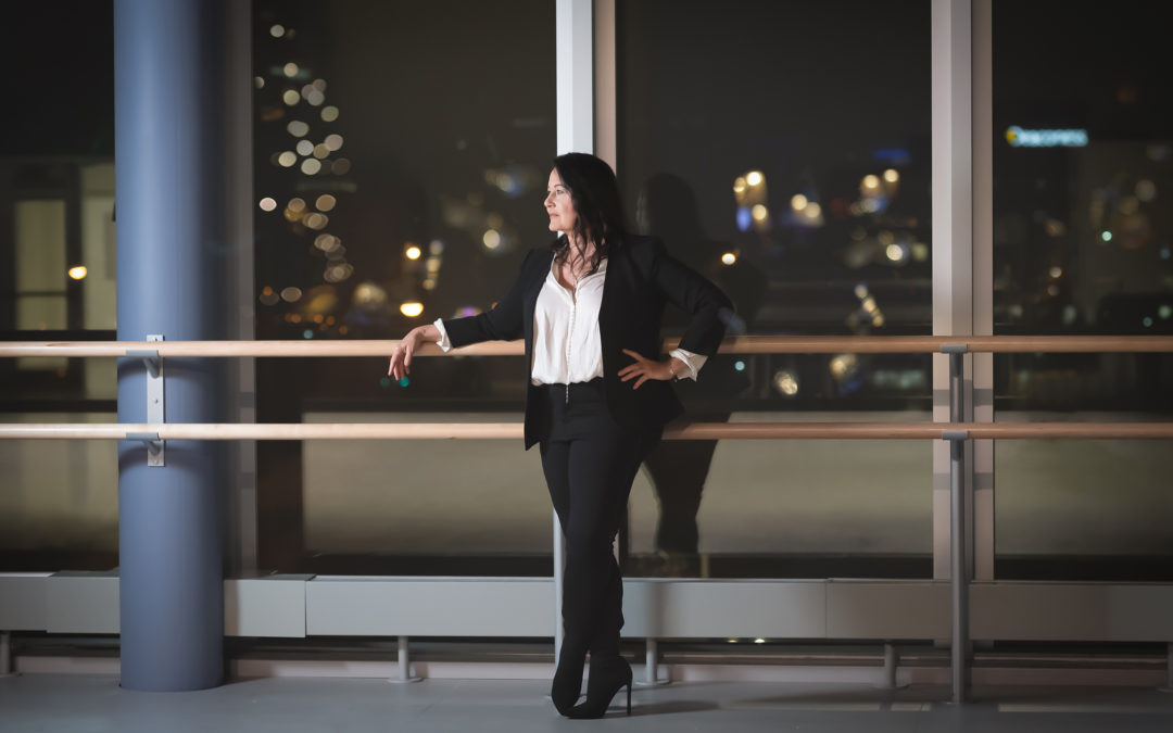 Jodie Gates, wearing a white blouse, dark blazer and pants and black heeled boots, stands against a ballet barre and looks off to her right. Behind her are large, floor-to-ceiling windwos that look out on the city of Cincinnati, where it is nighttime.