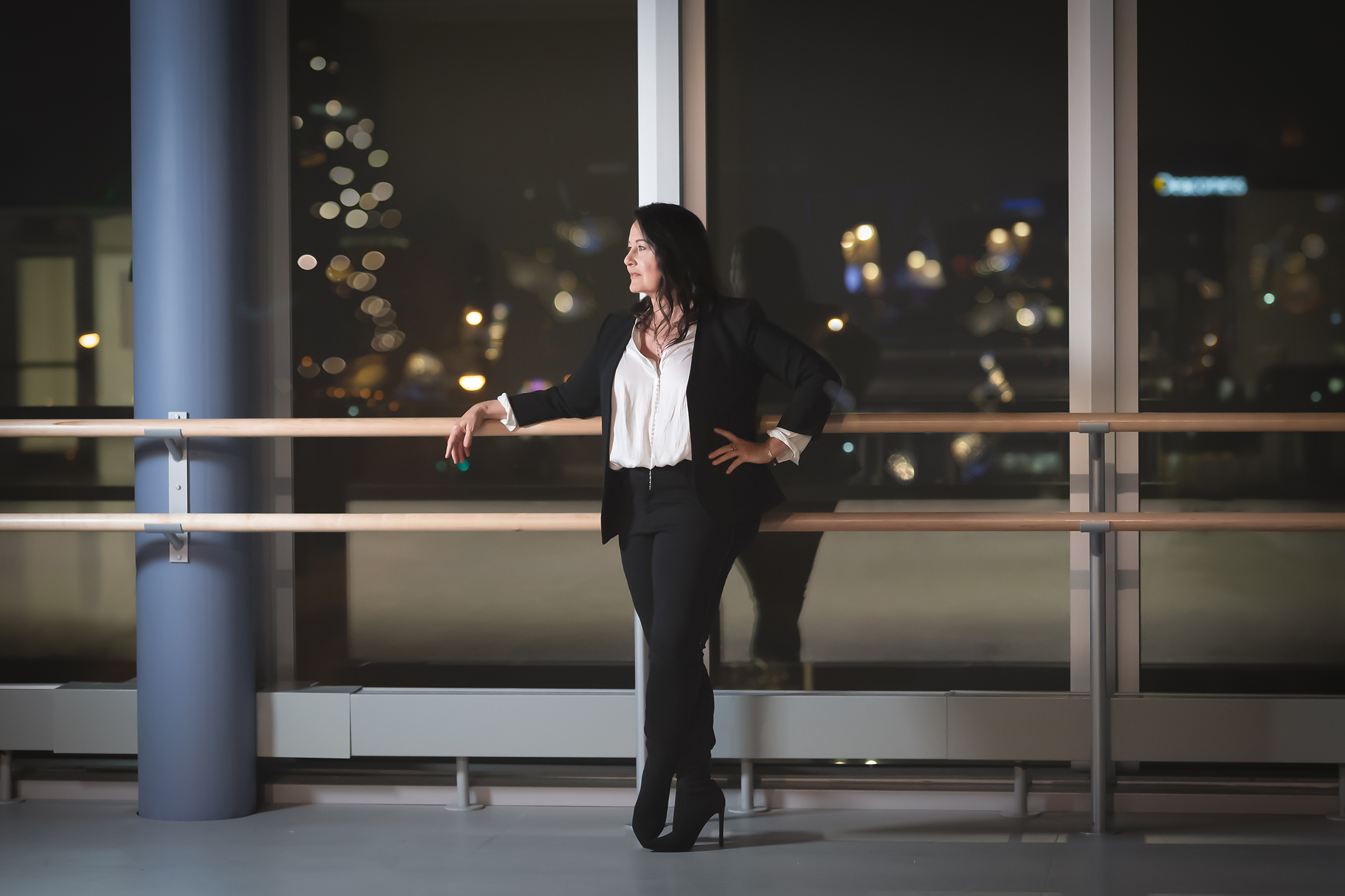 Jodie Gates, wearing a white blouse, dark blazer and pants and black heeled boots, stands against a ballet barre and looks off to her right. Behind her are large, floor-to-ceiling windwos that look out on the city of Cincinnati, where it is nighttime.