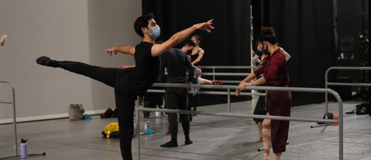 Ballet dancers taking class onstage with portable barres. In the foreground, Jonathan Montepara, in black dance clothing and a mask, balances in relevé arabesque.