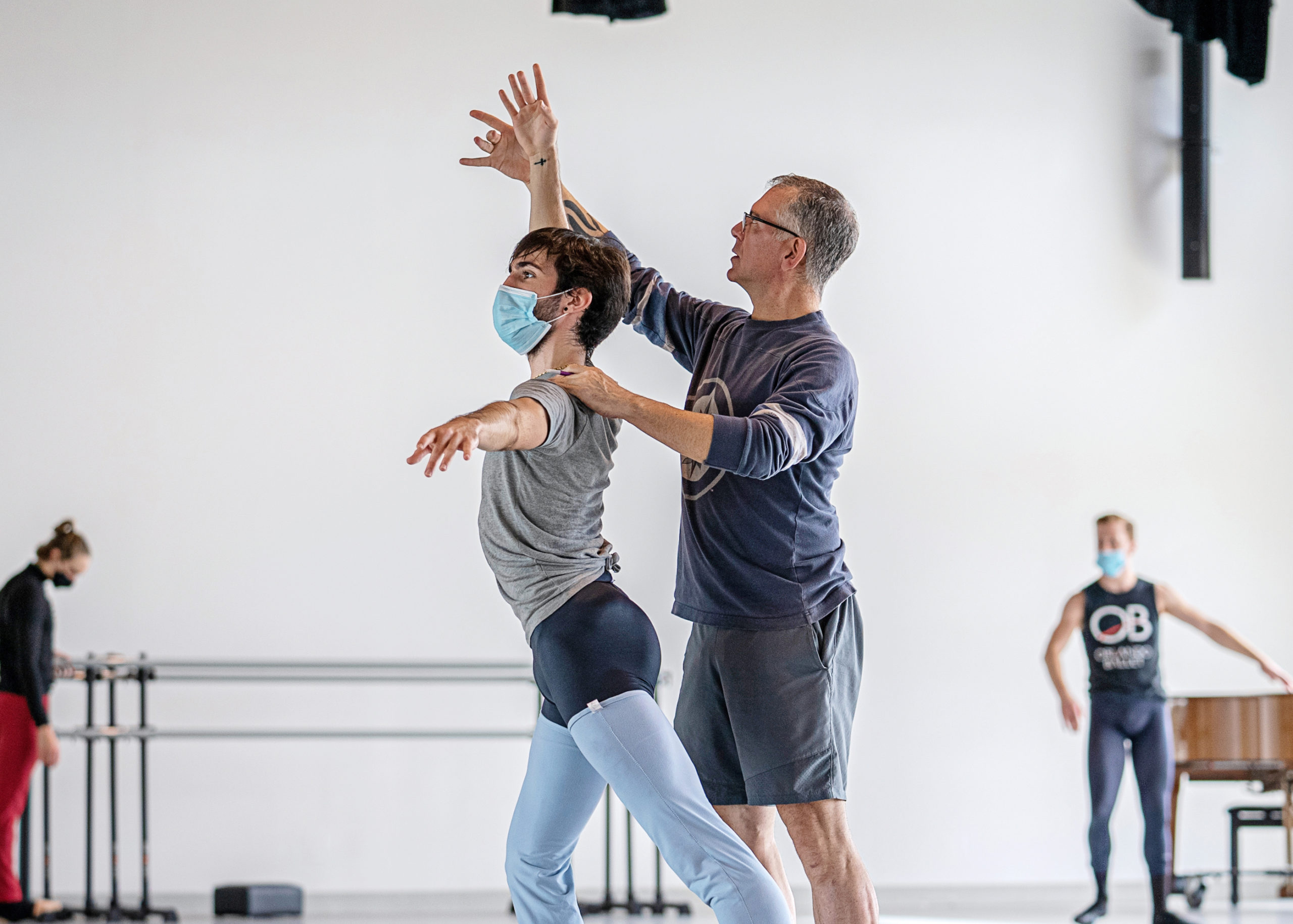 In a large dance studio, Jorden Morris, wearing a long-sleeved blue T-shirt and gray shorts, stands behind dancer Jaysan Stinnett and places his left hand on his shoulder while adjusting the dancer's raised right arm. Stinnett poses in an open fourth position and wears a face mask, gray T-shirt, black tights and blue, thigh-length leg warmers.