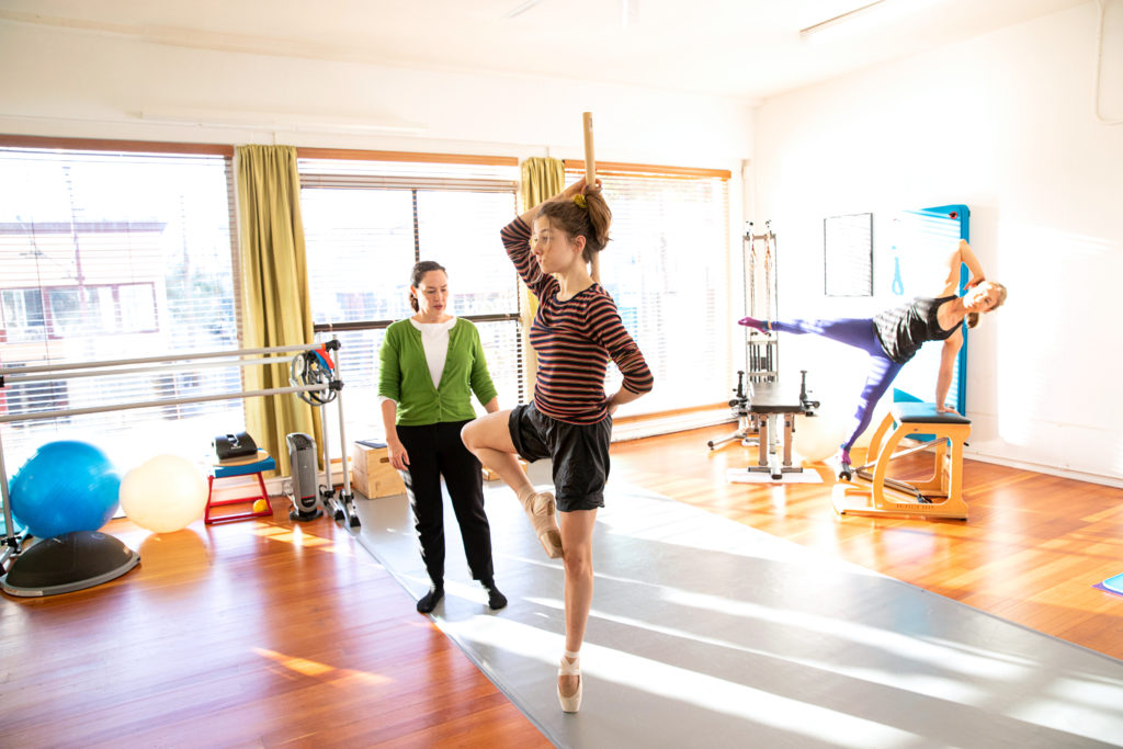 In a sunny physical therapy studio. Kendall Alway stands closely to a ballet dancer and watches as she performs a passé relevé on pointe. Alway wears a green cardigan over a white shirt and black sweatpants and socks while the dancer wears black shorts and a striped long-sleeved t-shirt. Behind them, another dancer in workout clothes practices an exercise on a Pilates chair.