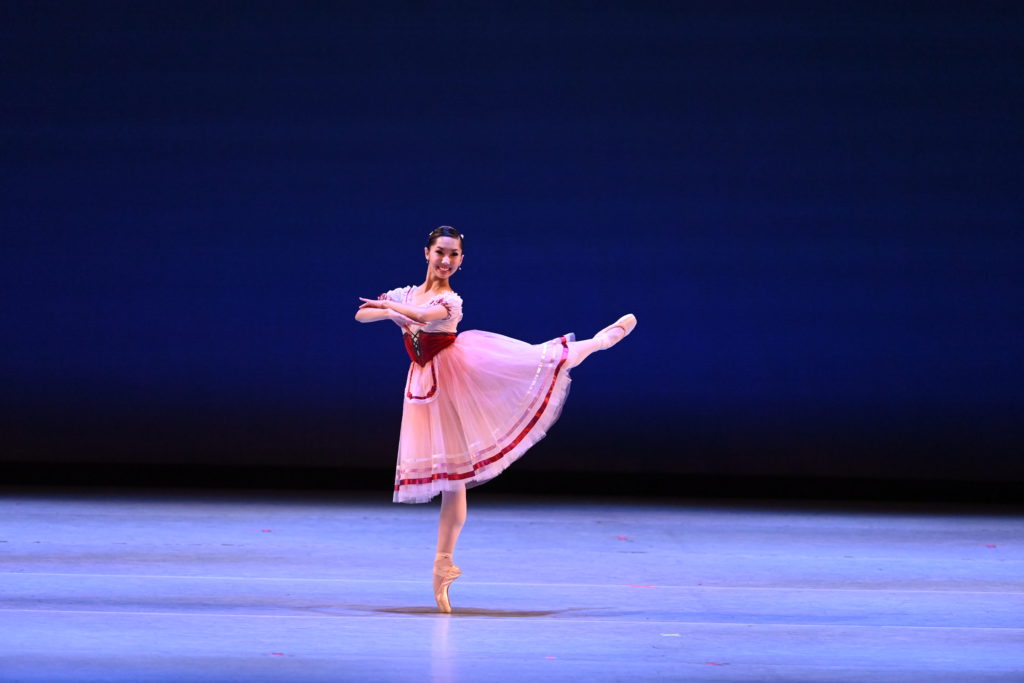 Lexi McCloud stands on a blank stage in an attitude derriere with her arms crossed at the elbows in front of her. She wears a pink Romantic-style tutu