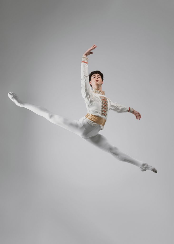 Mecah Levine jumps in a sissone en avan with his arms in fourth position. The background is blank behind him and he wears gray tights with a decorated long sleeve top.