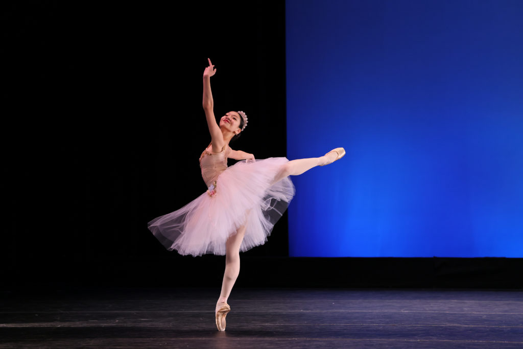 Michelle Lin poses in attitude derriere on a blank stage with her arms in an upturned fourth position. Her Romantic tutu swooshes around her legs; it is light pink.