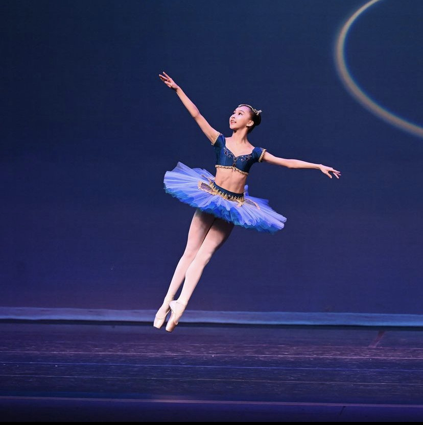 Nicole Liu jumps in an assemblé midair with her arms in open fourth. She wears a two-piece blue tutu.