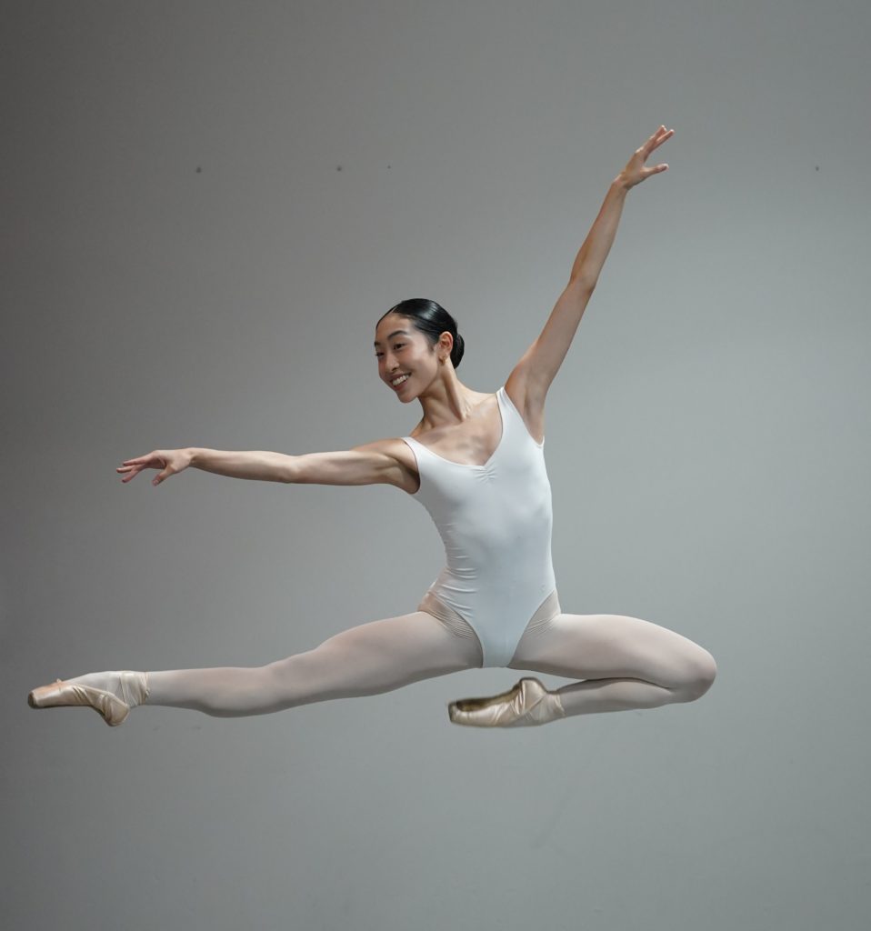 Cici Yu, a young Asian woman, is photographed midair with her right leg in second and her left tucked under her. She is wearing tights, pointe shoes and a light blue leotard.