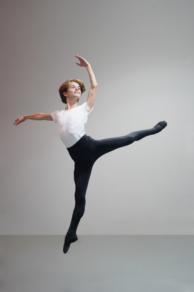 A young male dancer, in a white shirt and black tights, photographed in sauté middair with his back leg in attitude.