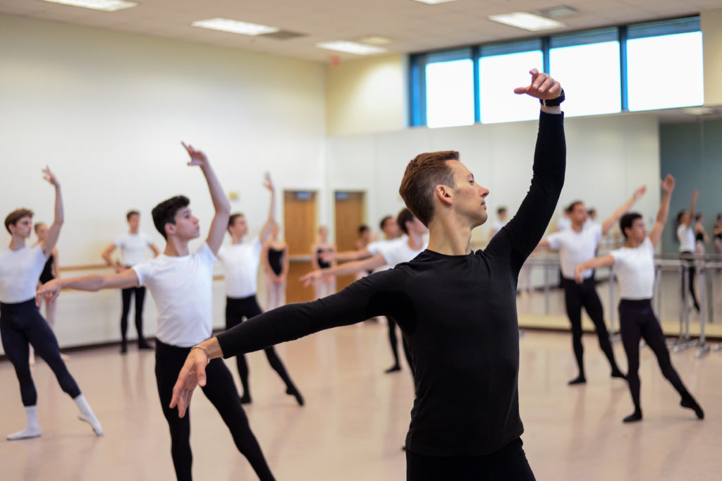 In a large dance studio, Philip Neal, wearing a black long-sleeved shirt, demonstrates his port de bras in ecarté with his left arm up. Behind him, a class of boy dancers wearing white T-shirts and black tights and ballet slippers follow him, doing tendu ecarté with their left leg extended.