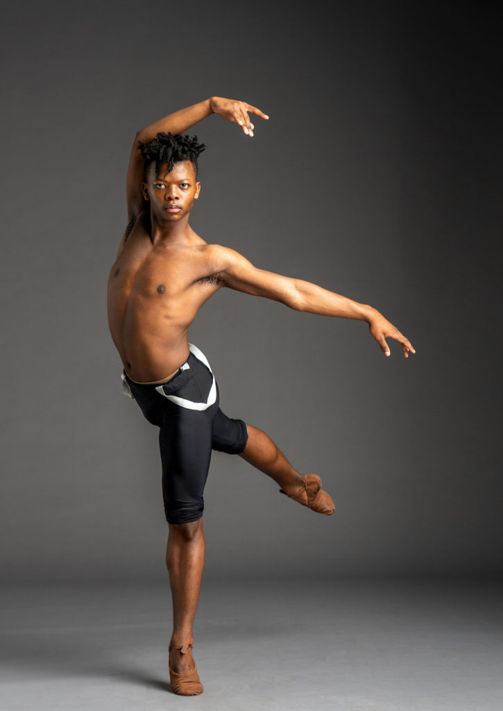Siphesihle November poses in front of a gray background in a low croisé arabesque on his left leg. He curves his right arm over his head and extends his left arm out to the side. He is barechested and wears black, knee-length tights and brown ballet slippers.