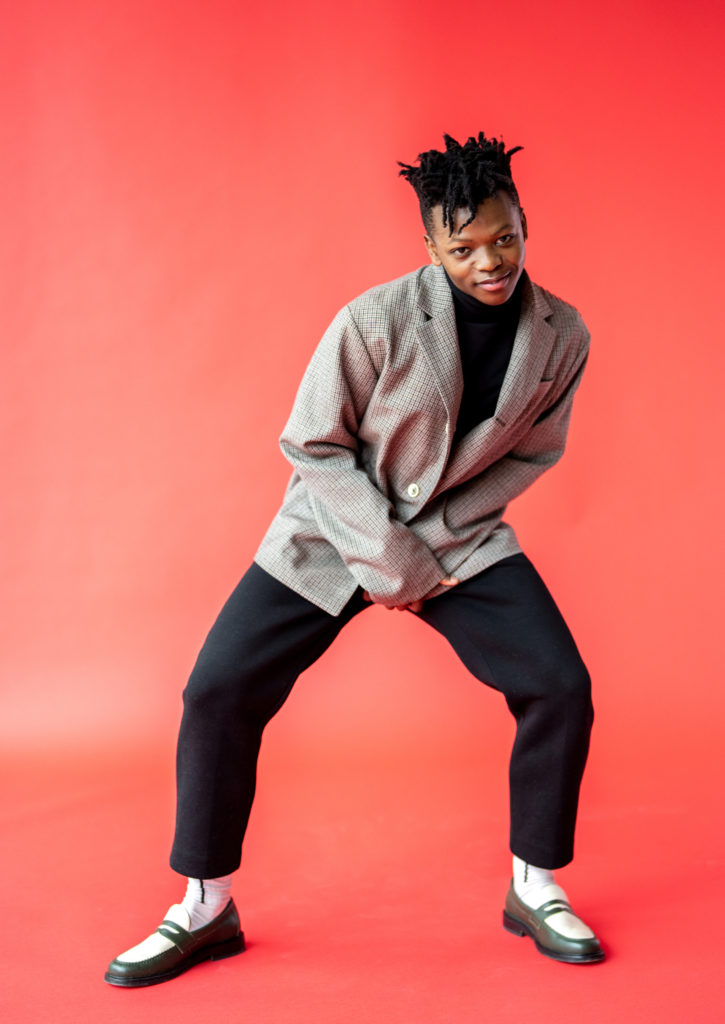 Siphesihle November poses in front of a hot pink backdrop with his legs in a wide stance, knees bent, and bending slightly forward fromt eh waist with a smile. He wears an oversized gray sportcoat, ankle-length black pants, white socks and white and green penny loafers.