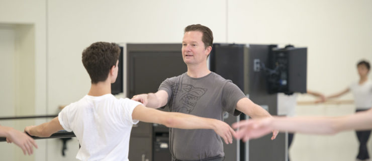 Peter Stark wears a gray T-shirt and pants and demonstrates a turn preparation to a class of teenage male dancers. The dancers face him in the studio and wear white T-shirts, black tights, white socks and ballet slippers.