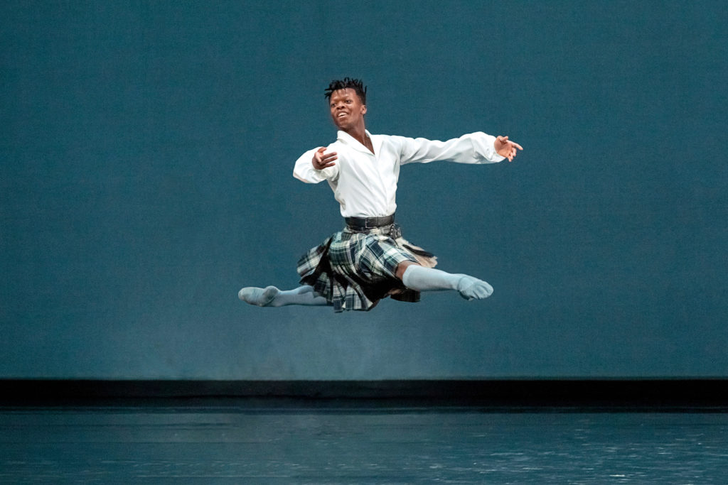 Siphesihle November performs onstage costumed in a plaid kilt, white long-sleeved shirt and gray knee-socks and ballet slippers. He is shown doing a Bournonville-style grand jeté en avant with his right leg in attitude derriere.